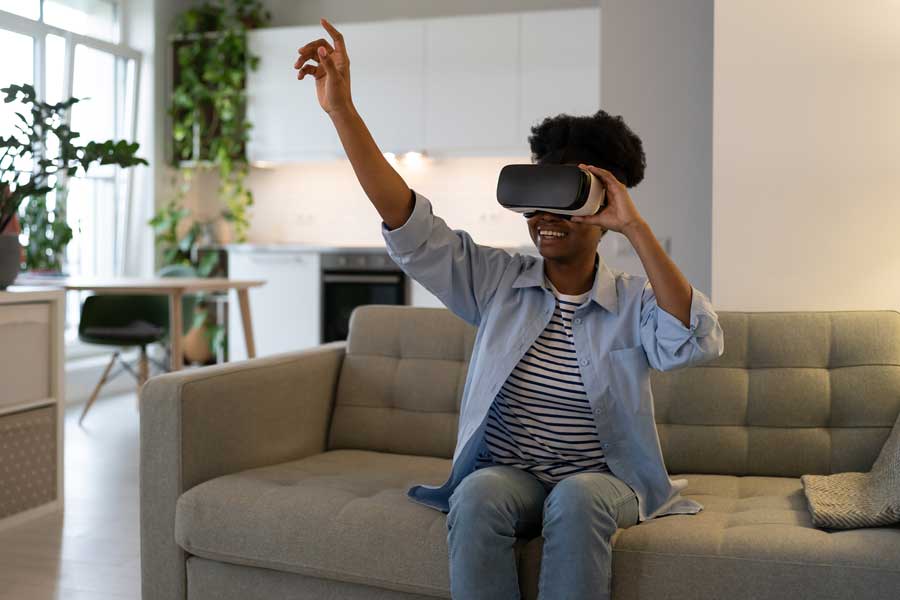 A woman sits on a couch with a VR headset on. She smiles and reaches up a hand.