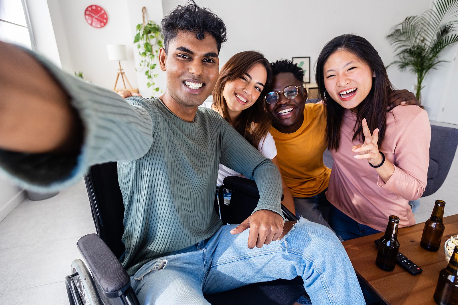 Inclusion and diversity concept with young indian man in wheelchair taking selfie portrait with multiracial friends. United group of diverse young people having fun together at home