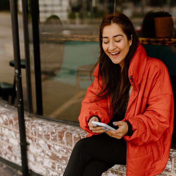 a woman in an orange jacket sitting on a window sill smiling excitedly at her phone
