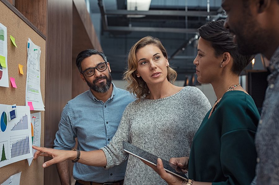 A group of diverse business professionals engaged in a collaborative meeting, discussing charts and data on a bulletin board. The image highlights the importance of face-to-face networking, personal connections, and accountability in local networking groups such as BNI.