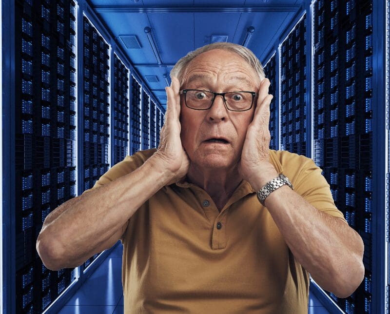 Older man with glasses in yellow polo shirt holding head in shock inside data center with illuminated server racks