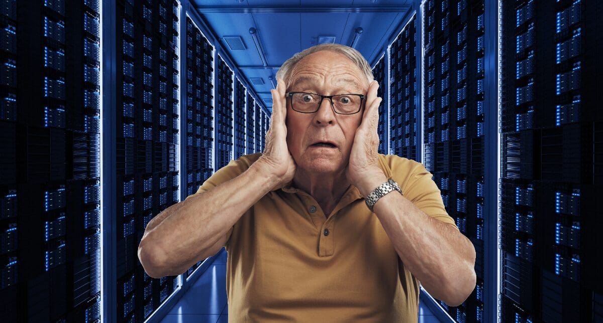 Older man with glasses in yellow polo shirt holding head in shock inside data center with illuminated server racks